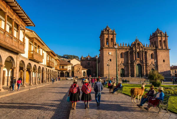 Menschen gehen und sitzen rund um die Plaza de Armas in Cusco, Peru, mit kolonialen Gebäuden und einer historischen Kirche bei klarem blauem Himmel.
