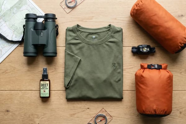 Draufsicht auf Ausrüstung für Wildlife-Abenteuer mit grünen Ferngläsern, einem gefalteten olivgrünen T-Shirt, Insektenschutzspray, einem Kompass, einer orangefarbenen Trockenpacktasche, einem orangenen Schlafsack und einer schwarzen Stirnlampe auf einer Holzoberfläche.
