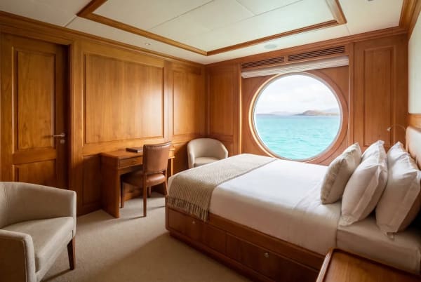 Gemütliche Kreuzfahrtschiffkabine mit Holzverkleidung, großem rundem Fenster mit Meerblick, Bett mit weißen Bettwäsche und Kissen, zwei Sesseln und einem kleinen Schreibtisch mit Stuhl.