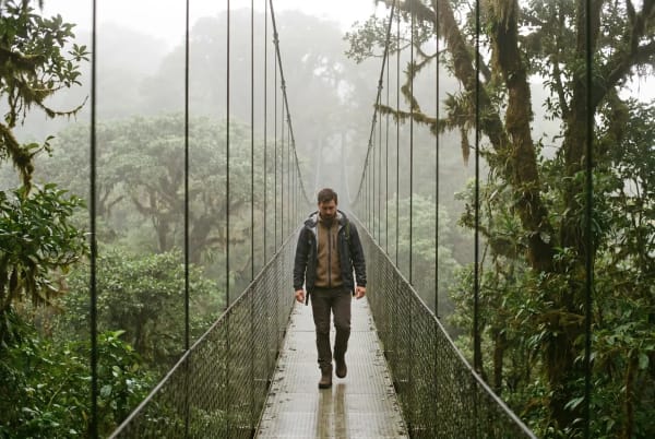 Ein Mann läuft allein auf einer schmalen Hängebrücke, umgeben von dichtem, nebligem Wald mit üppigen grünen Bäumen.