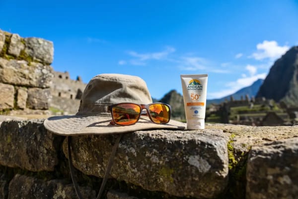 Sonnencreme mit Lichtschutzfaktor 50 und ein breitkrempiger Hut mit Sonnenbrille liegen auf einer alten Steinmauer vor bergiger Kulisse und blauem Himmel.