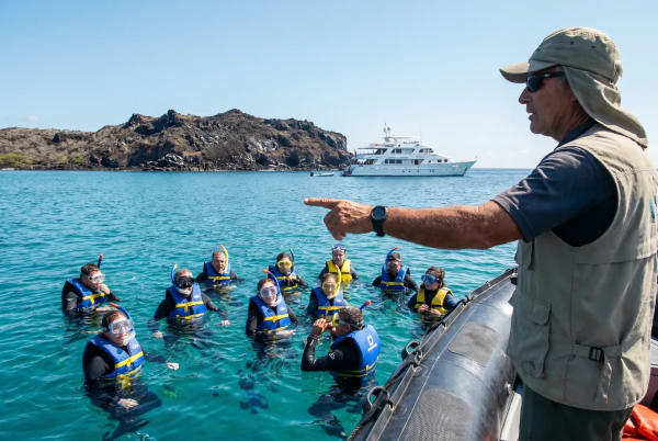 Eine Gruppe von Schnorchlern mit Schwimmwesten, die im klaren blauen Wasser nahe einer felsigen Insel schwimmen, während ein Führer auf einem Boot auf sie zeigt und im Hintergrund eine große weiße Yacht liegt.