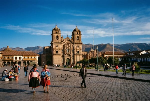 Menschen gehen und versammeln sich auf einem sonnigen Platz vor einer historischen Kathedrale mit Bergen im Hintergrund in Cusco, Peru.