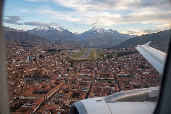 Blick auf die Stadt Cusco mit ziegelgedeckten Dächern und einer Landebahn in der Mitte, umgeben von Bergen mit schneebedeckten Gipfeln, gesehen aus einem Flugzeugfenster während des Landeanflugs.