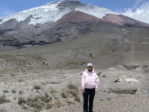 Eine Person mit hellrosa Kapuzenpullover und lila Mütze steht in einer kargen, felsigen Landschaft mit einem schneebedeckten Berg im Hintergrund unter einem teilweise bewölkten Himmel.