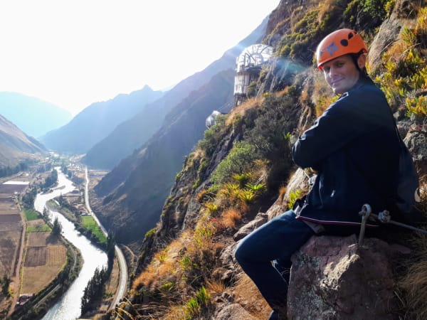 Person mit orangefarbenem Helm und dunkler Jacke sitzt am Rand eines felsigen Abhangs mit Blick auf das Heilige Tal in Peru, darunter ein gewundener Fluss und Berge im Hintergrund.
