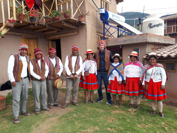 Gruppe von Männern und Frauen in traditioneller peruanischer Kleidung steht vor einem rustikalen Gebäude mit Pflanzen und Wassertanks im Hintergrund.