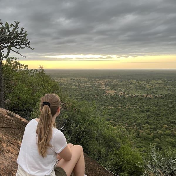 Eine Frau mit langen blondem Haar in einem Zopf, trägt ein weißes T-Shirt, Shorts und Turnschuhe, sitzt auf einem felsigen Hügel und blickt auf eine weite Landschaft mit bewölktem Himmel bei Sonnenuntergang.