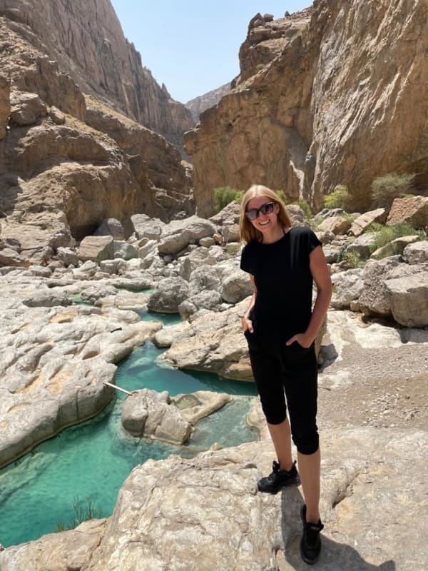 A woman standing on a rocky outcrop in a deep canyon with turquoise water flowing through it, surrounded by tall cliffs.