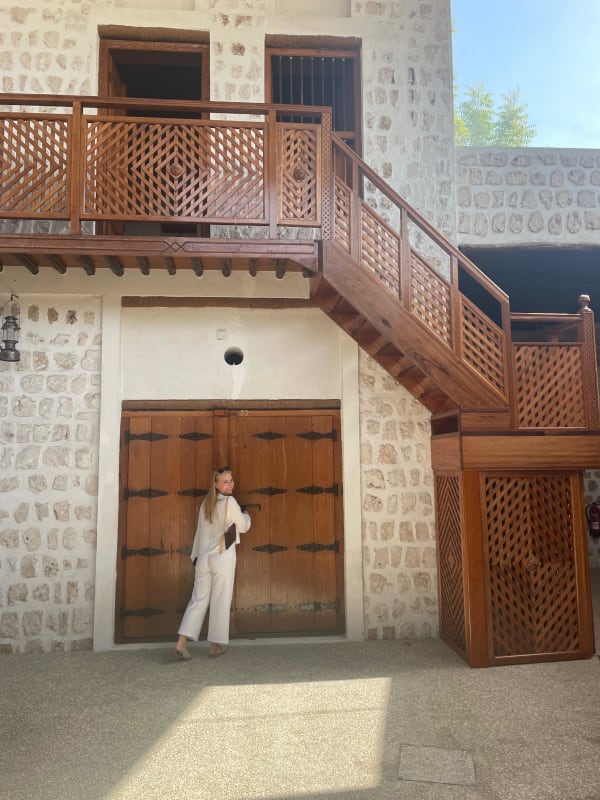 A woman in white pants and a white top stands near large wooden doors in a stone building with a balcony and staircase.
