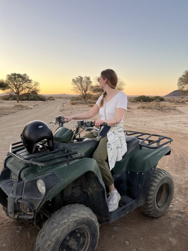 A woman in casual clothing sitting on a quad bike in a desert landscape at sunset.