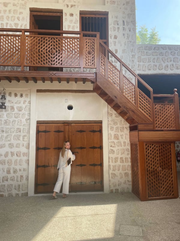 A woman in beige pants and a white top stands in front of a large wooden door, with a stone wall and wooden staircase behind her.