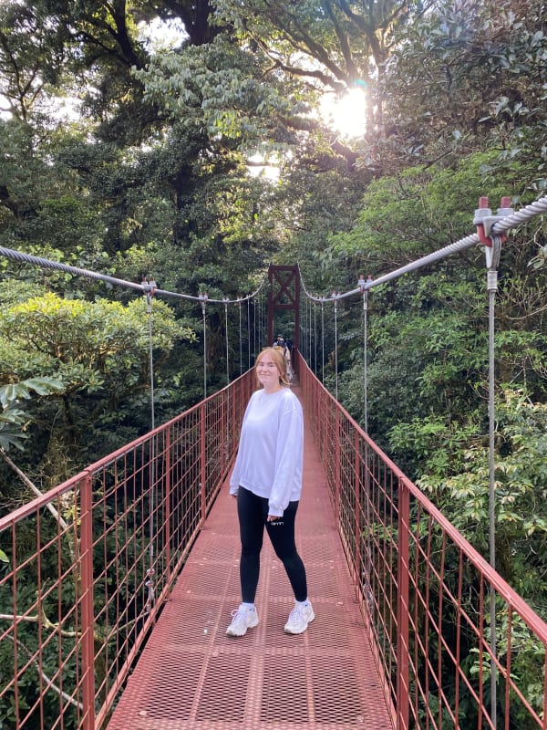 Eine junge Frau steht auf einer roten Hängebrücke, umgeben von dichtem grünen Dschungel mit Sonnenlicht, das durch die Bäume scheint.