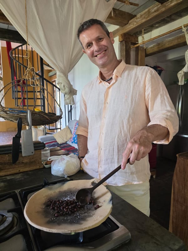 Ein lächelnder Mann in einem hellbeigen Hemd röstet Beeren auf einer traditionellen Tonschale über einem Gaskocher in einer rustikalen Kücheneinrichtung mit Holzbalken und einer Wendeltreppe im Hintergrund.
