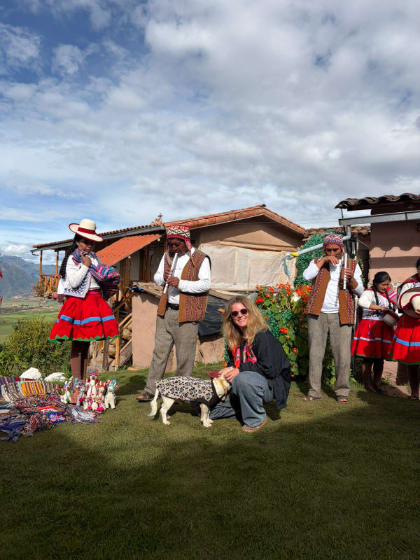 Eine Gruppe von Menschen in traditioneller peruanischer Kleidung steht auf Gras neben handgefertigten Textilien und Figuren, im Hintergrund sind Berge und Häuser zu sehen. Eine Frau mit Sonnenbrille hockt sich hin und streichelt einen kleinen Hund mit Pullover.