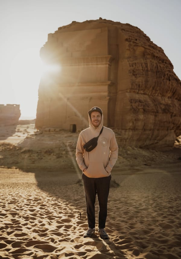 A man standing on sand with ancient ruins in the background during sunset.