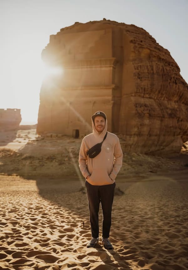 A smiling man standing on sand with an ancient stone structure and the sun setting behind it in Saudi Arabia.