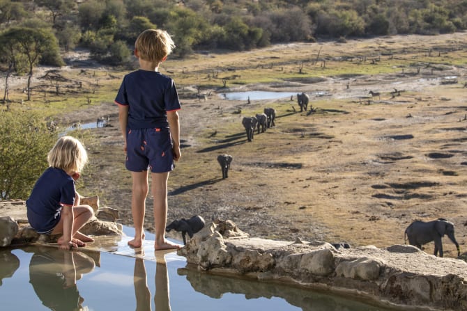 Zwei Kinder stehen am Rand eines Pools und blicken auf eine weite, trockene Savanne, in der eine Gruppe von Elefanten zu einem Wasserloch zieht.