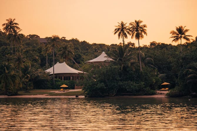 A tropical lakeside scene at sunset with lush greenery, palm trees, and thatched-roof buildings.