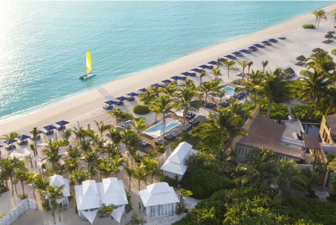 An aerial view of a beach resort with lounge chairs, umbrellas, and palm trees along a sandy shoreline, with a sailboat in the water.