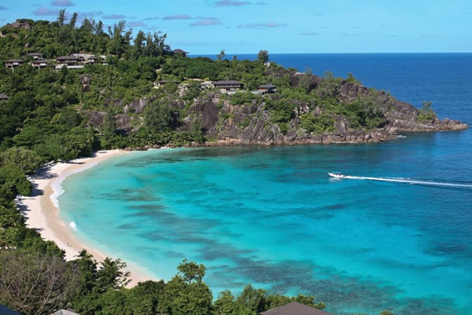 Ein malerischer Blick auf einen halbmondförmigen Strand mit türkisfarbenem Wasser, üppigem Grün und Resort-Gebäuden auf einer tropischen Insel.