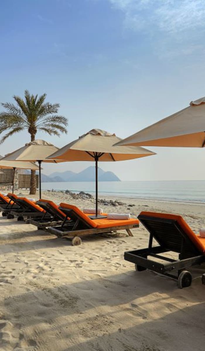 Eine Reihe leerer Liegestühle mit orangefarbenen Kissen und großen Sonnenschirmen am sandigen Strand, mit Palmen und Bergen im Hintergrund.