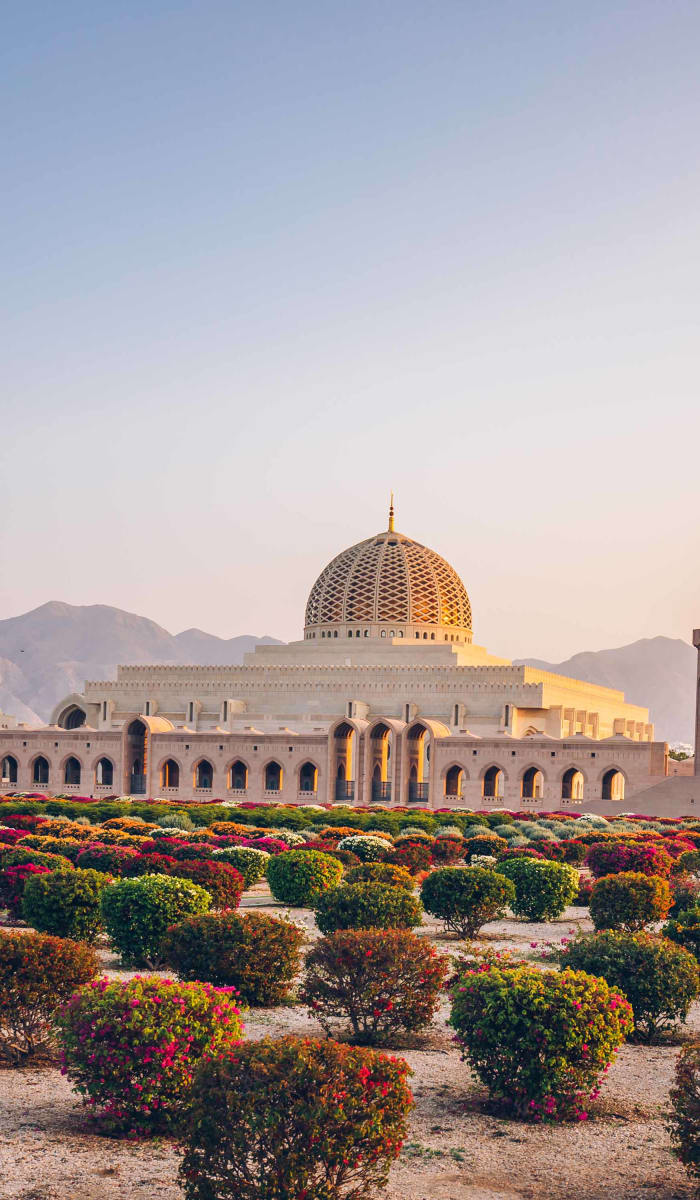 A mosque with a large central dome and four minarets situated behind a well-maintained garden with evenly spaced shrubs, set against a mountainous backdrop at sunset.