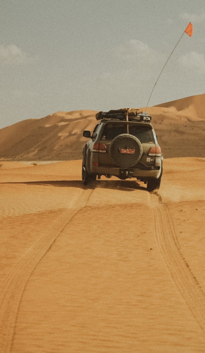 Ein Jeep fährt durch eine sandige Wüstenlandschaft mit Dünen und bewölktem Himmel im Hintergrund.