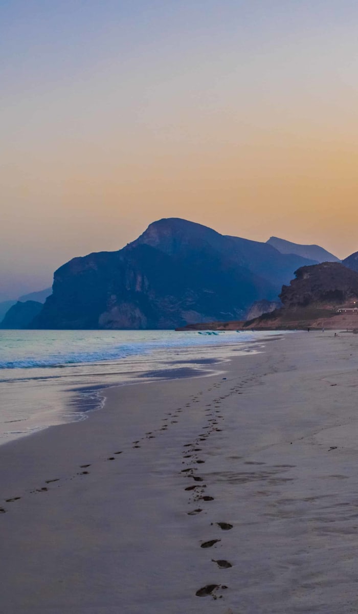 A tranquil beach scene at sunset with footprints leading along the sandy shore and mountains in the background.