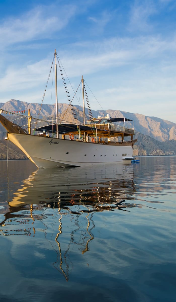 A white traditional dhow boat floating on calm waters with mountainous landscape in the background.