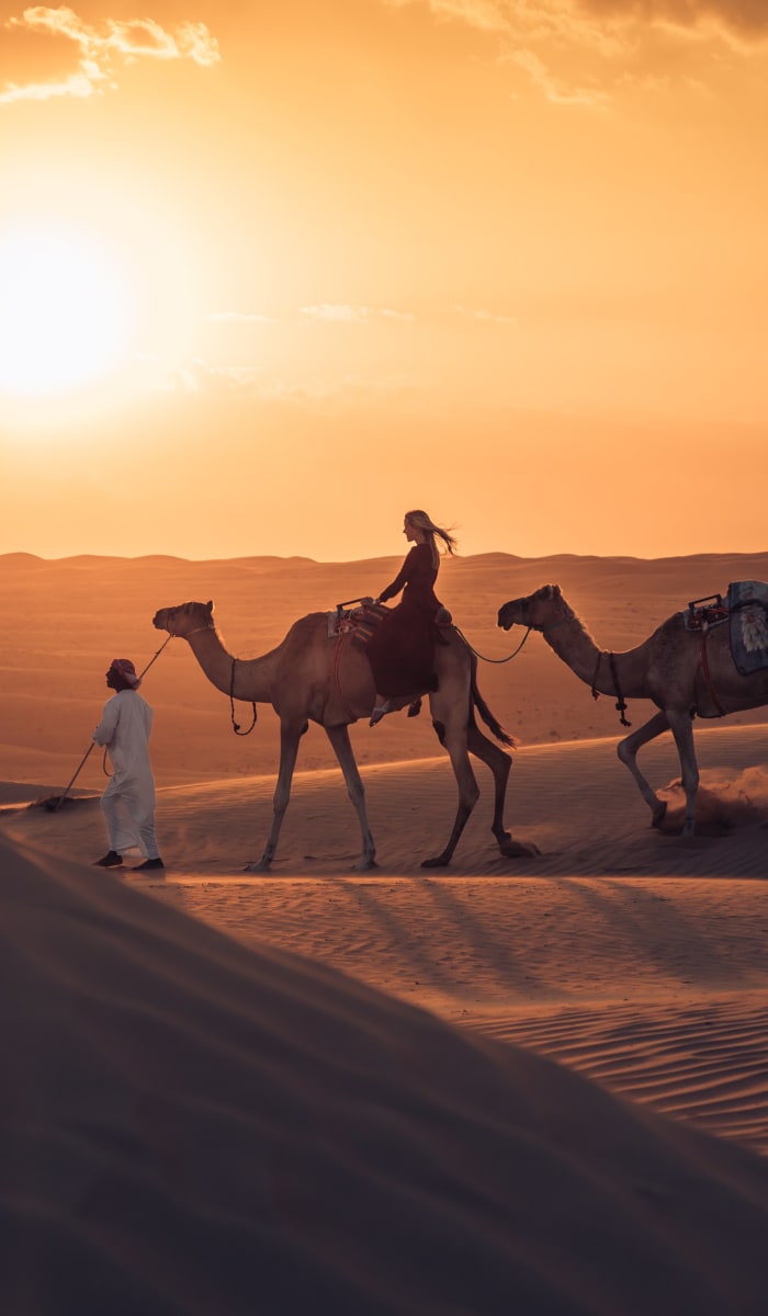 A person leading two camels with riders through the desert at sunset in the Oman Wahiba Desert.