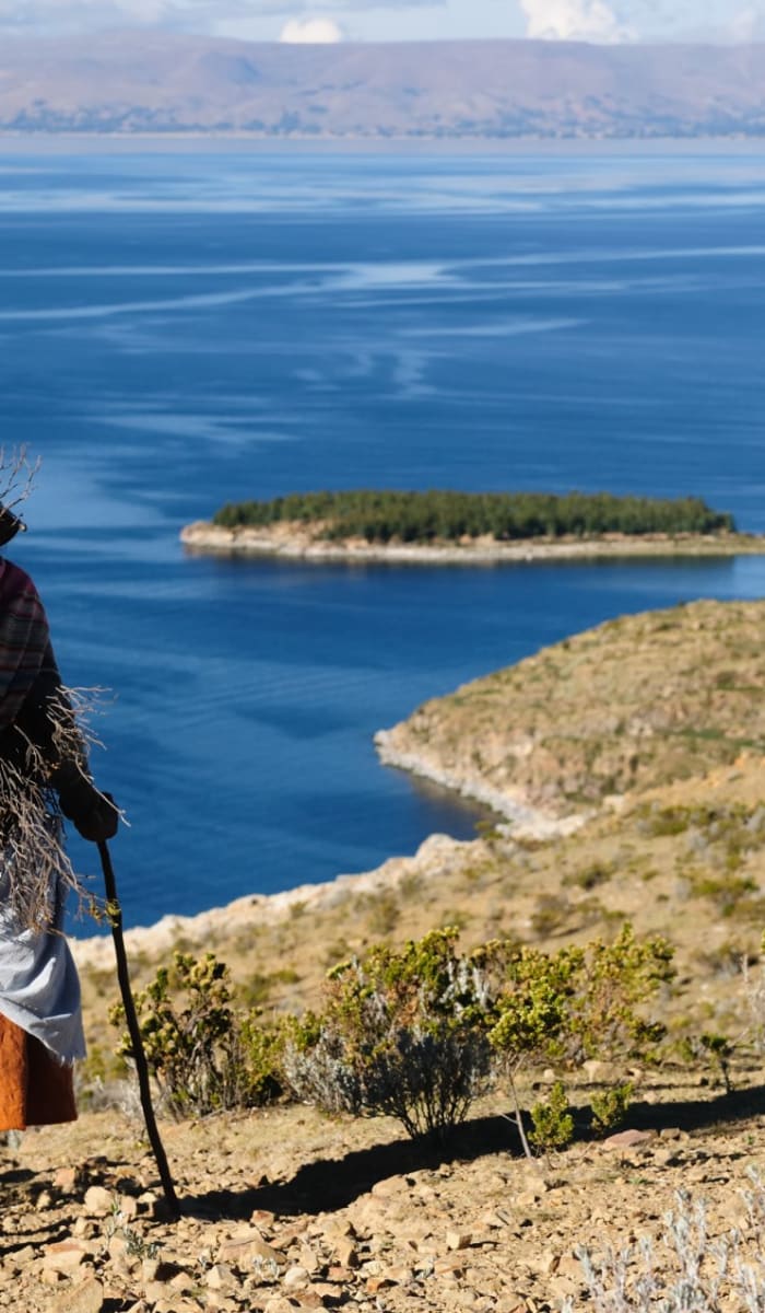 Elderly woman in traditional clothing stands on a hillside overlooking Lake Titicaca with islands in the distance.