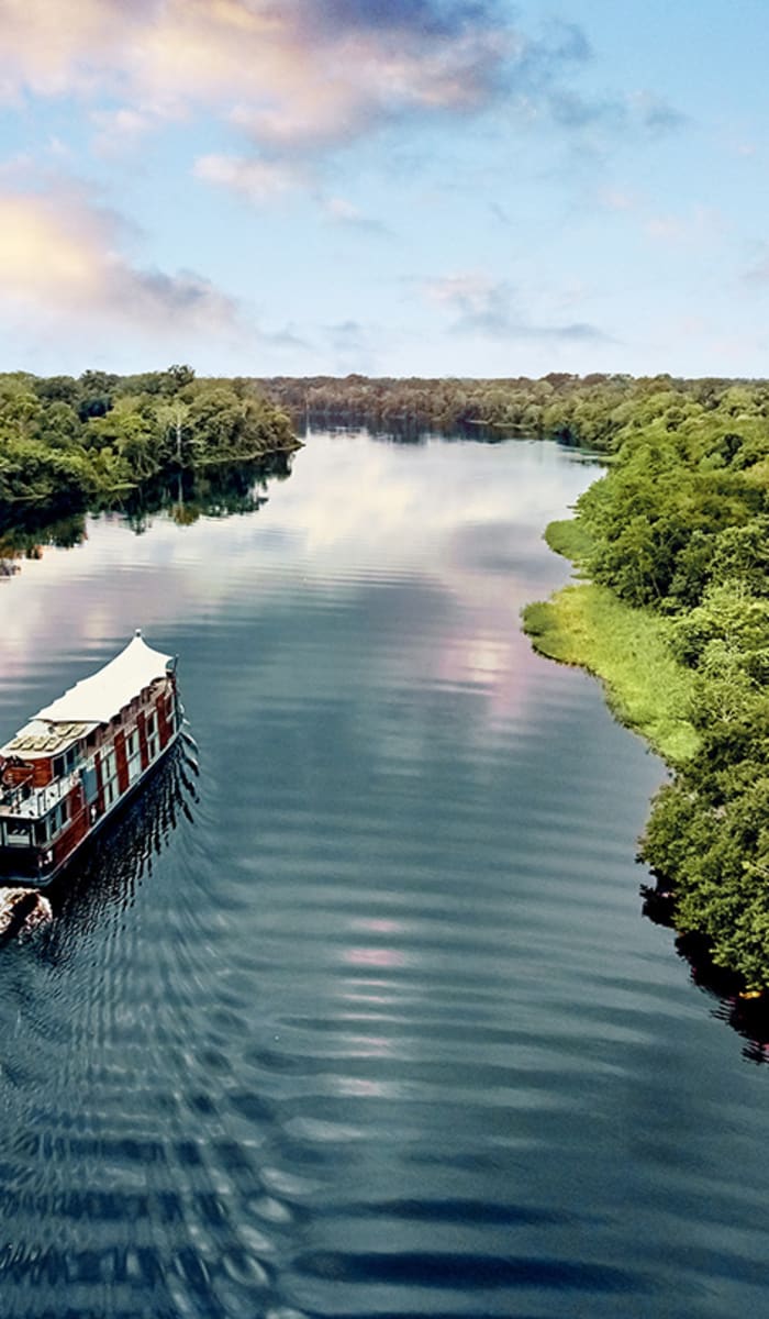 A boat sailing on a wide river surrounded by dense green rainforest under a partly cloudy sky.