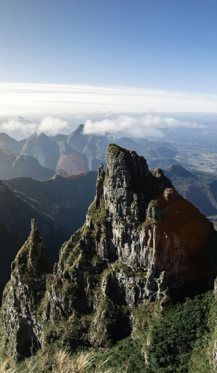 Rocky mountain cliffs covered with some vegetation under a bright, clear sky with distant misty peaks and valleys.