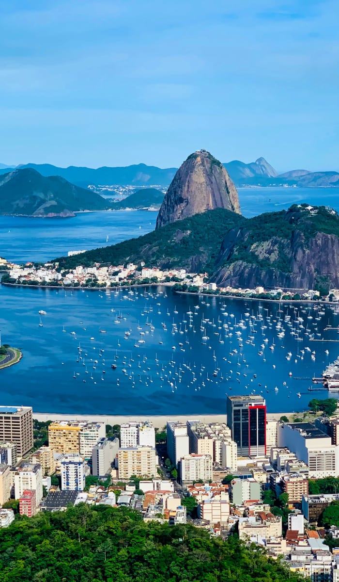 A panoramic view of Rio de Janeiro featuring Sugarloaf Mountain rising behind a bay filled with numerous boats and surrounded by urban buildings and green hills.