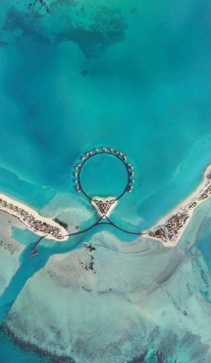 Luftaufnahme eines luxuriösen Resorts mit kreisförmigen Wasserbungalows, die durch eine Brücke verbunden sind, umgeben von türkisfarbenem Wasser und Sandstränden.
