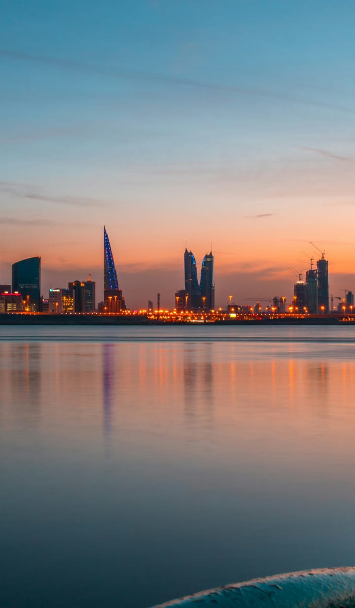 A city skyline during sunset reflected on a calm body of water with rocks in the foreground.