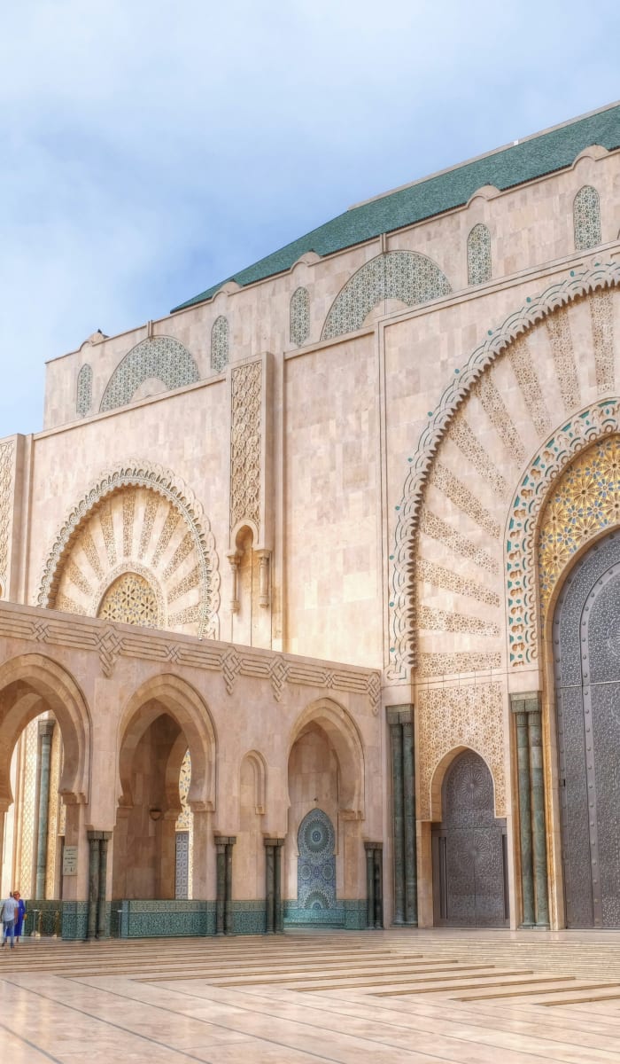 Der große verzierte Eingang einer Moschee in Casablanca mit detaillierten Bögen und dekorativen Fliesen, mit wenigen Menschen, die draußen auf dem Boden sitzen.