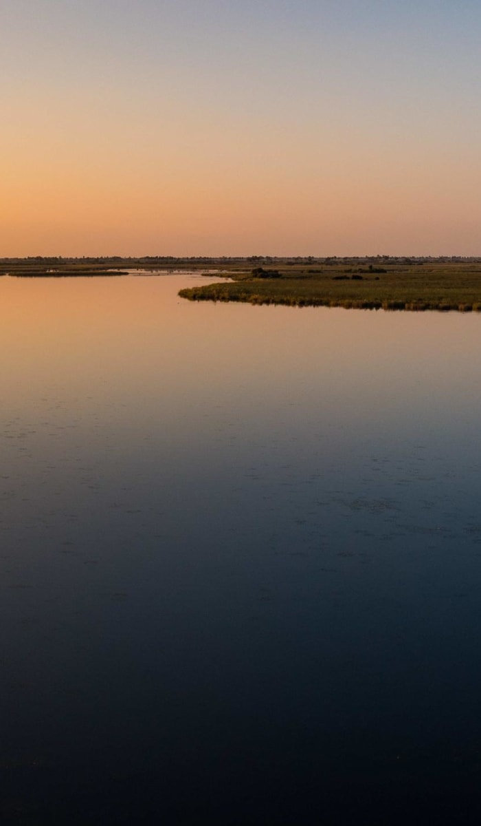 Ein Wasserfahrzeug fährt bei Sonnenuntergang über einen weiten, ruhigen Fluss mit offenen Ebenen im Hintergrund.