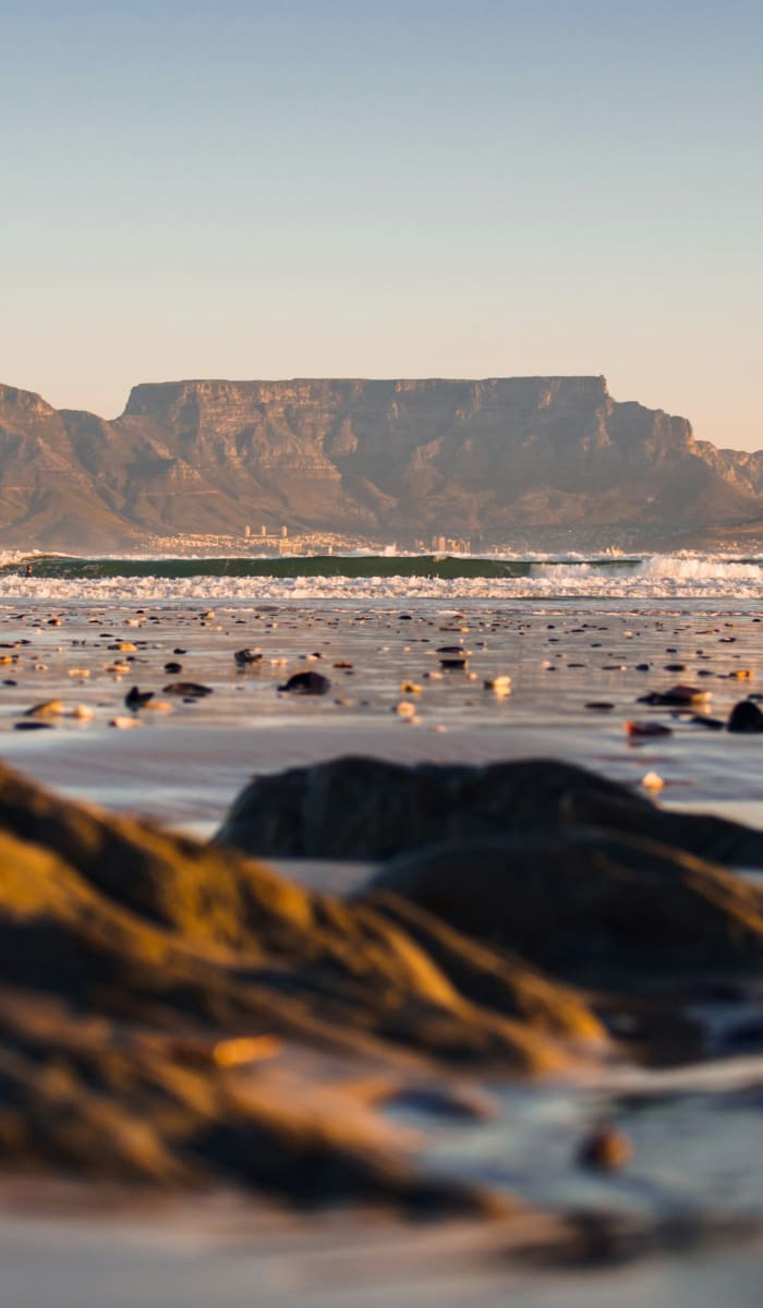 Blick auf den Tafelberg von einem Strand aus, mit Felsen und nassem Sand im Vordergrund während des Sonnenuntergangs.