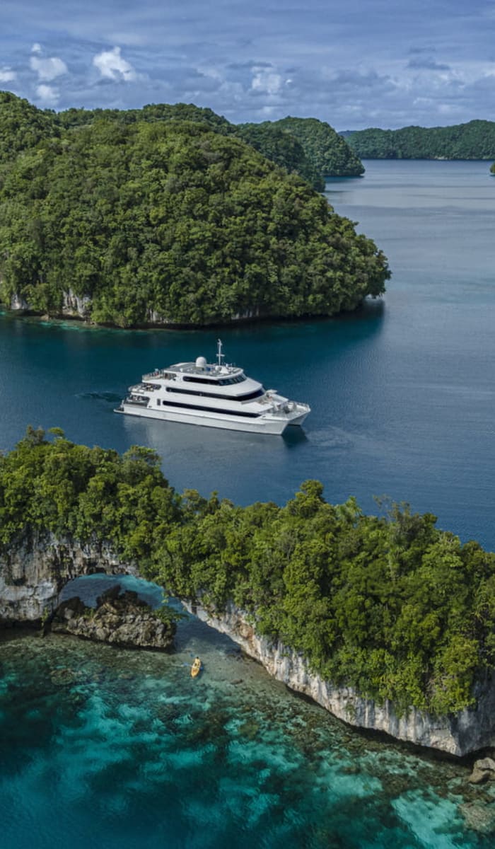 Eine Luxusyacht vor üppig grünen Inseln in einem ruhigen Meer, mit klarem blauem Wasser und partly Wolken am Himmel.
