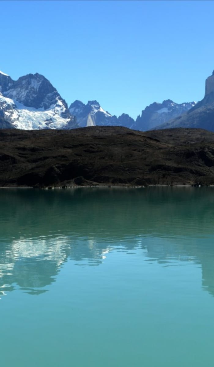 A panoramic view of snow-capped mountains reflected in a calm lake, set against a clear blue sky.