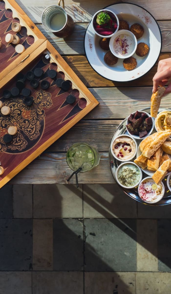 A top-down view of a table with traditional Lebanese food, a backgammon game, and a person reaching for food.