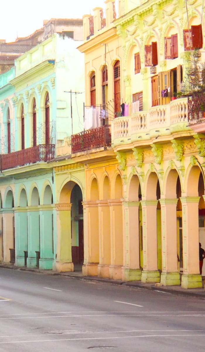 Colorful pastel buildings line a street with a vintage blue car driving by in Havana, Cuba.
