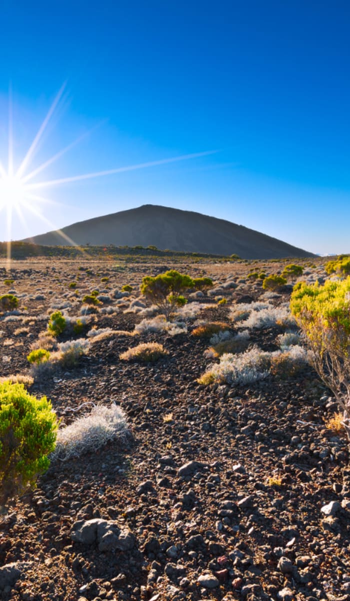 Bright sun shining over a rocky volcanic landscape with scattered green bushes and a mountain silhouette in the background under a clear blue sky.