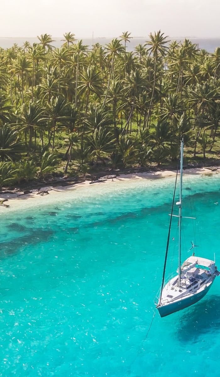 A sailboat floating near a tropical island with a dense palm tree forest and turquoise waters.