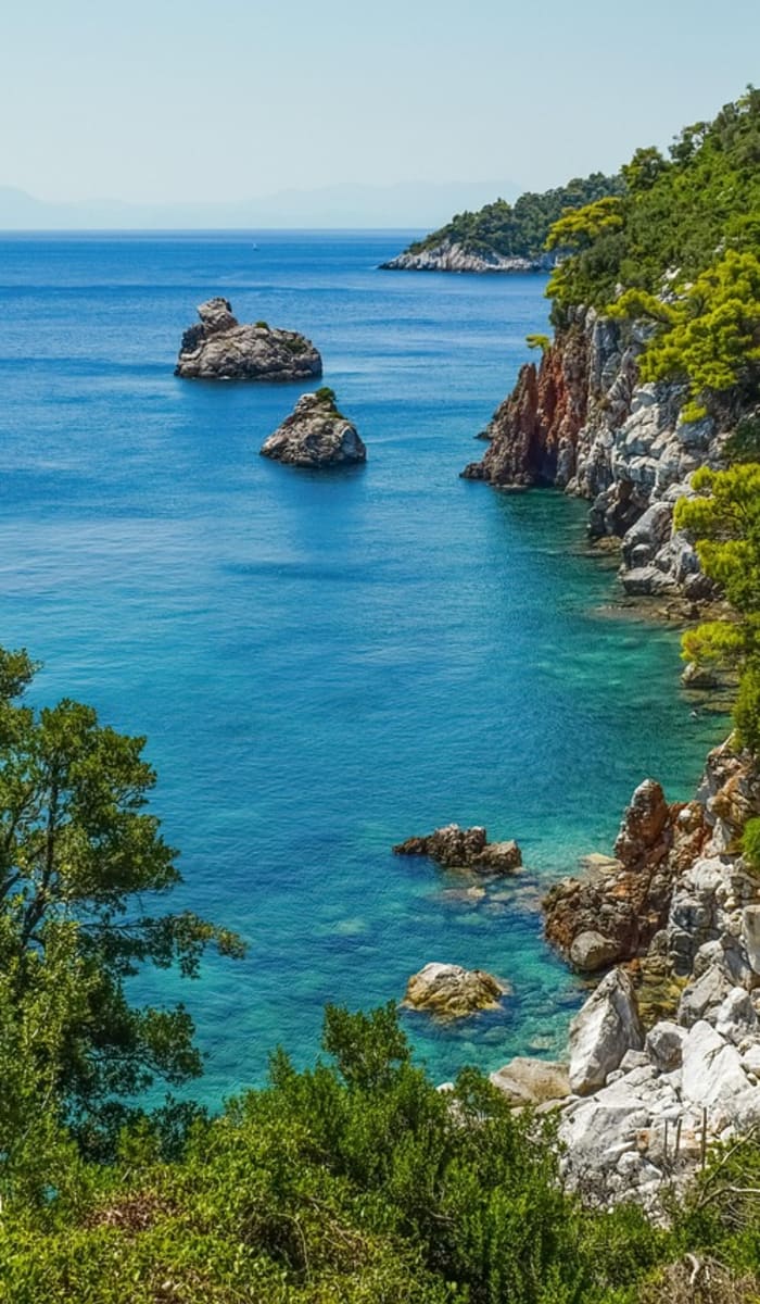 Küstenbucht von Alonissos mit klarem blauem Meer, felsigen Klippen und dichtem grünem Kiefernwald unter klarem Himmel.