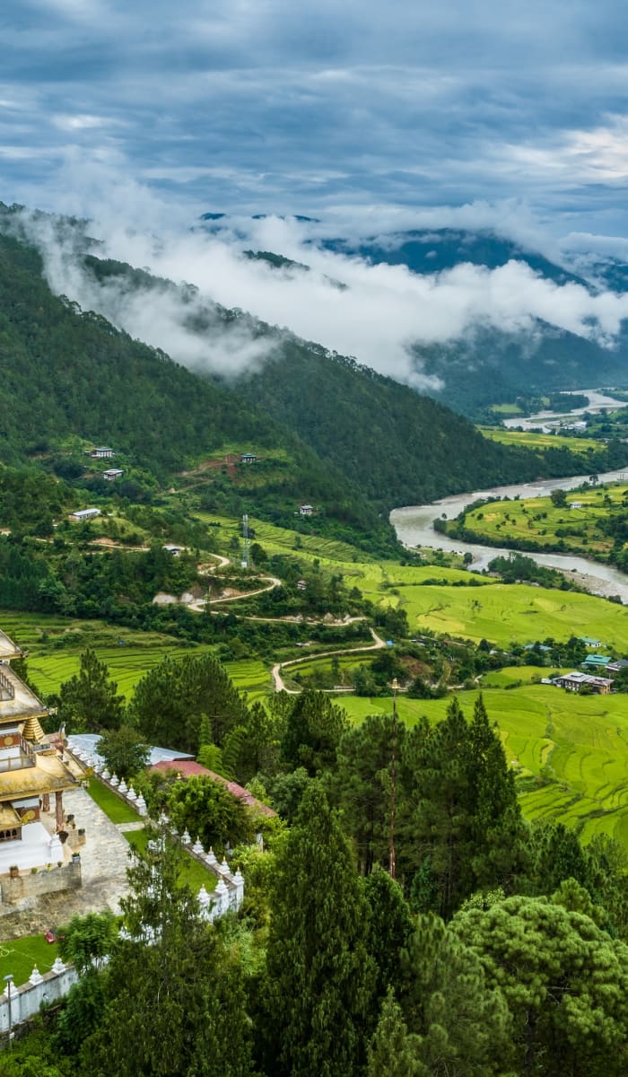 Ein buddhistisches Kloster gelegen inmitten üppiger grüner Bäume in einem Tal, mit einem Fluss, der durch die Landschaft mäandert, und nebelbedeckten Bergen im Hintergrund.