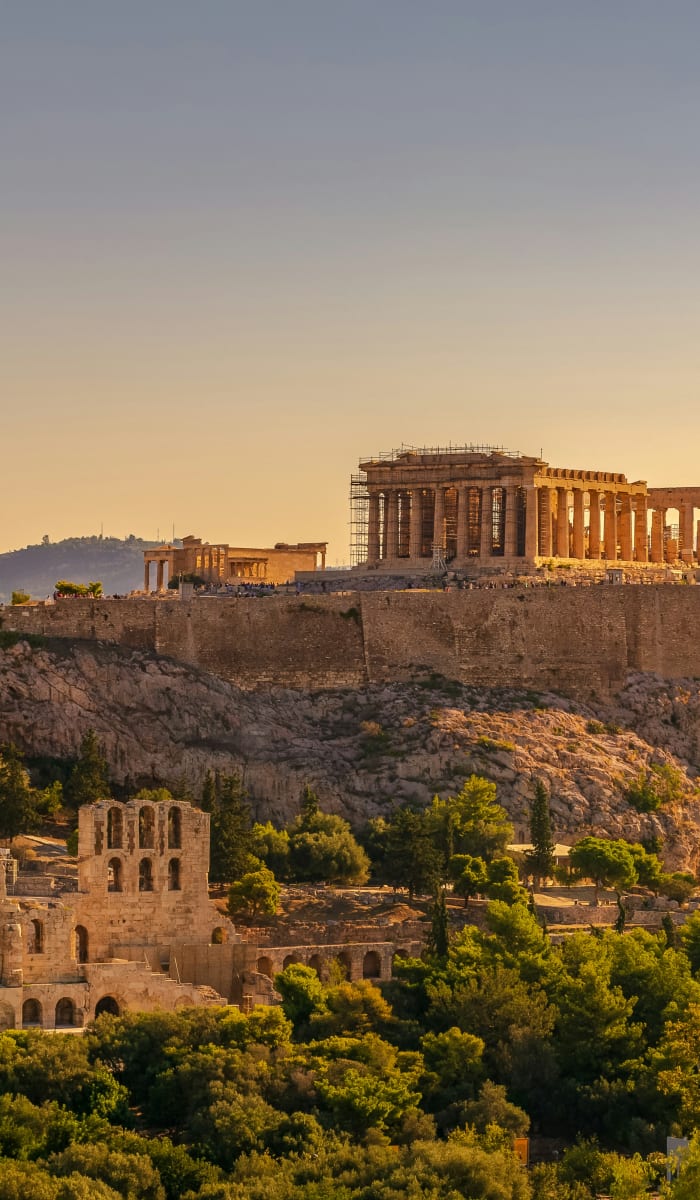 Die Akropolis von Athen auf einem felsigen Hügel bei Sonnenuntergang, mit dem antiken Parthenon-Tempel und umgeben von Grün und weiteren historischen Ruinen.