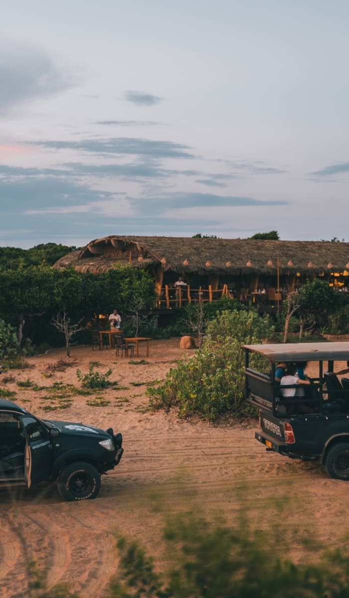 Zwei schwarze Safari-Fahrzeuge geparkt auf einer staubigen Freifläche in der Nähe von üppigem Grünland und traditionellen Hütten bei Sonnenuntergang.